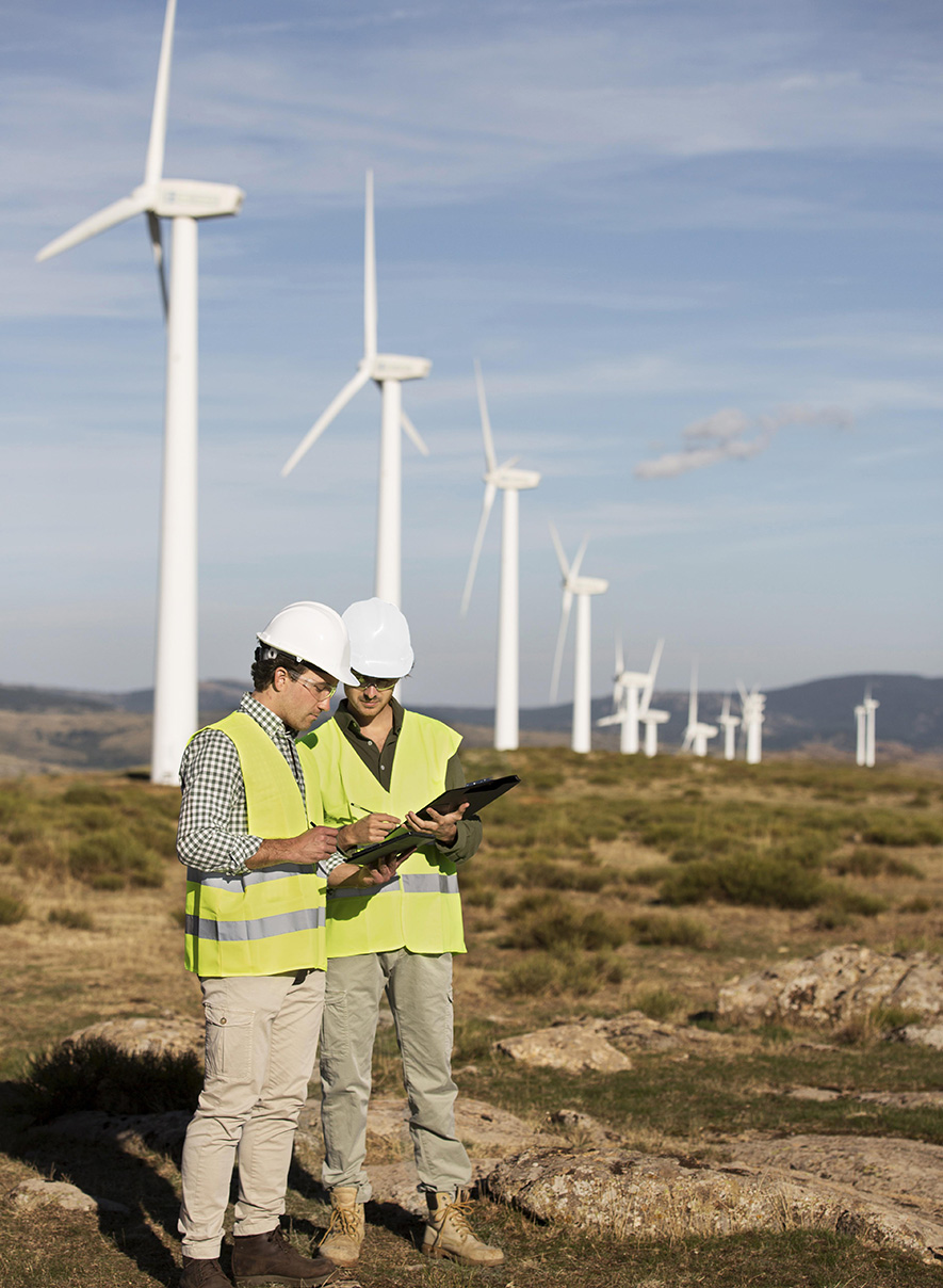 Two engineers in high-visibility vests and hard hats review a tablet at a wind farm with row of turbines in the background.